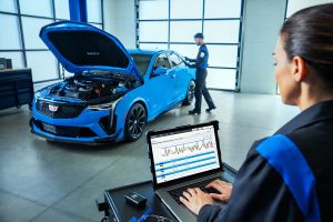 A mechanic working on a Cadillac vehicle, with the hood open, while another technician monitors diagnostic data on a laptop.